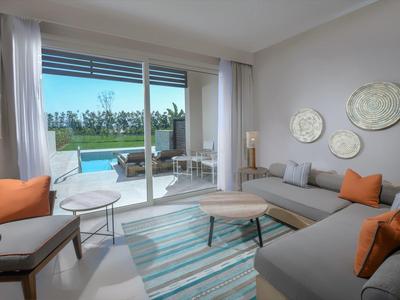 Moderner Wohnraum mit großen Fenstern, grauem Sofa, orangenen Kissen und Zugang zur Terrasse mit Pool.