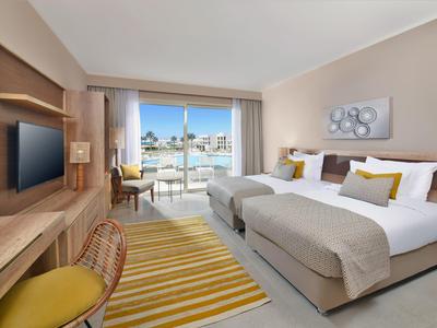 Helles Hotelzimmer mit zwei Betten, großem Balkon, Holzmöbeln und gelb-weißen Akzenten.