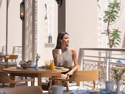 Frau sitzt in hellem Café mit Holztischen und Stühlen, modernes Ambiente, Tageslicht, Pflanze im Hintergrund.