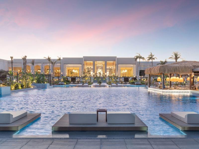 Luxuriöser Pool mit Sonnenliegen und Palmen vor großem, modernem Hotelgebäude bei Sonnenuntergang.