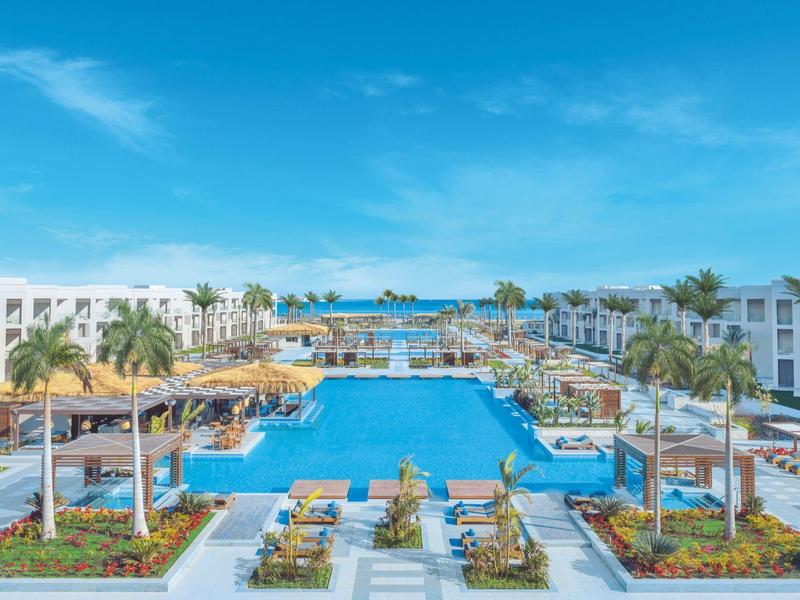 Großer blauer Pool mit Palmen, Sonnenliegen und weißer Hotelanlage unter blauem Himmel.