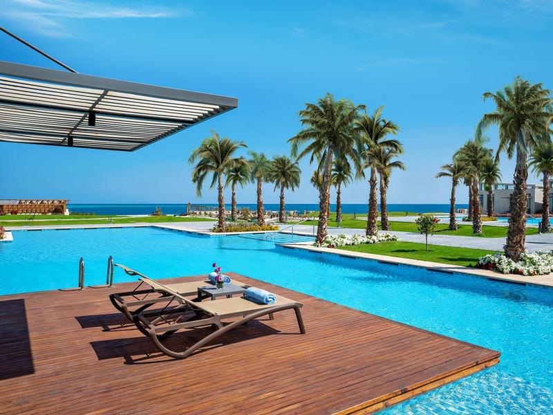 Pool area with lounge chairs, palm trees, and ocean view at a hotel resort.