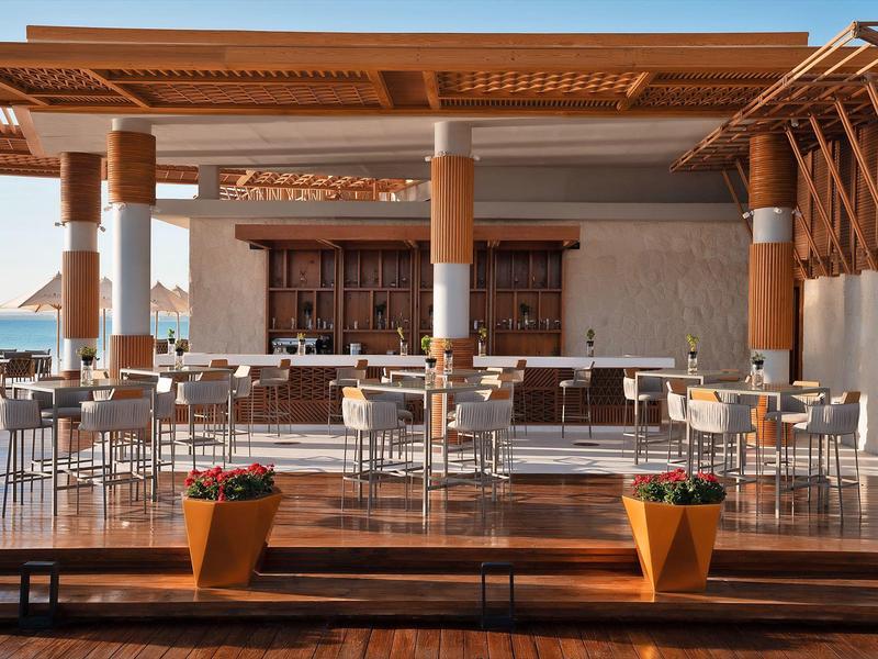 Modern outdoor hotel bar area with wooden columns and stools under blue sky.