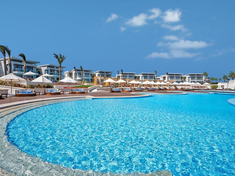 Large outdoor pool in front of a beach resort with sun umbrellas and blue sky.