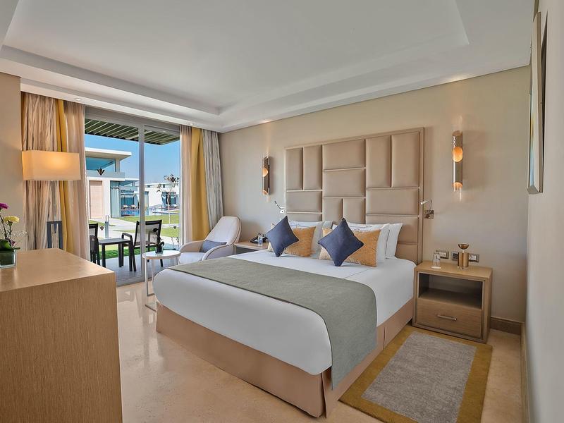 Modern hotel room with large bed, balcony, and view of beach bungalows.