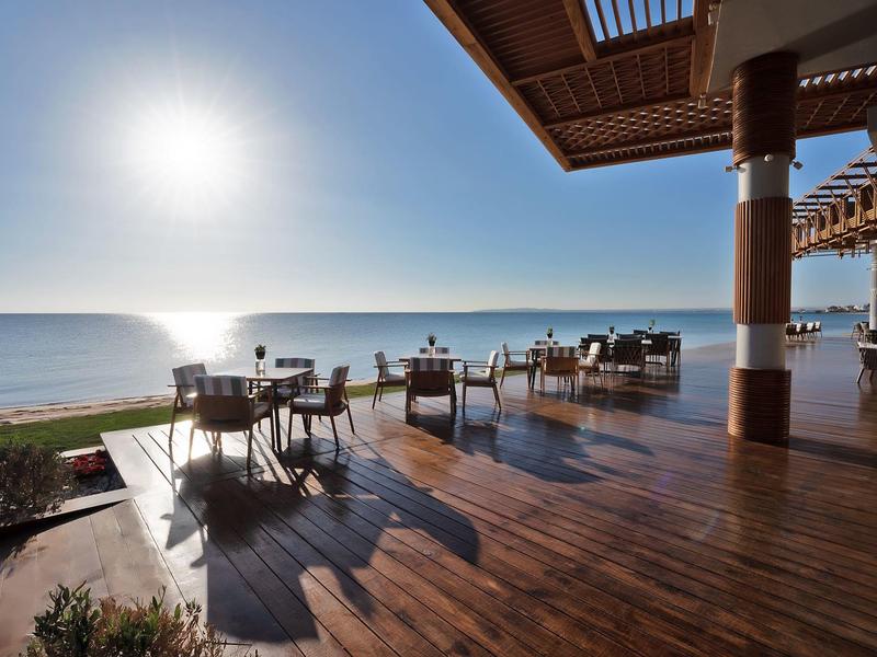 Open terrace with tables and chairs, view of beach and sea in sunny weather.