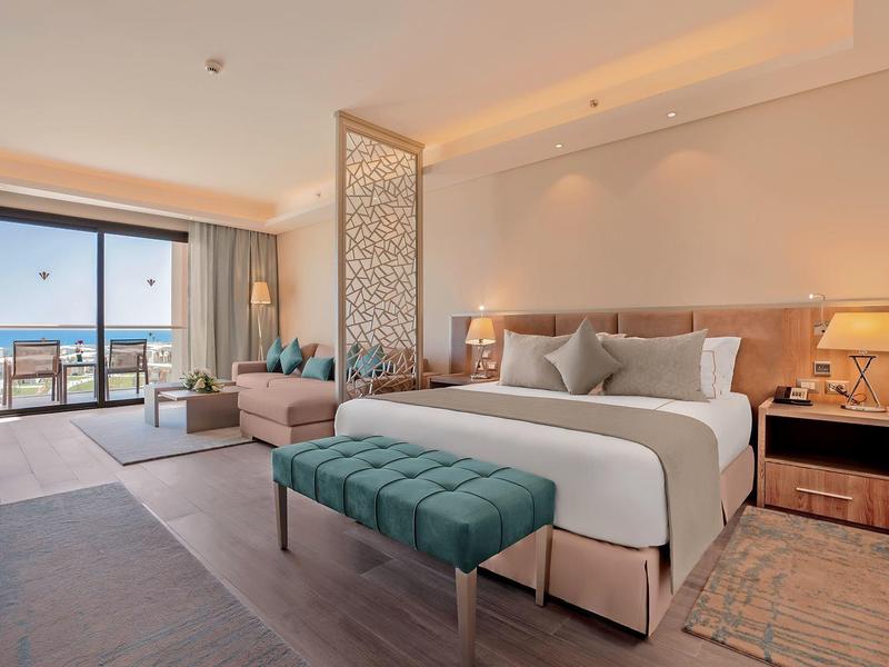 Modern hotel room with large bed, seating area, and balcony with sea view.