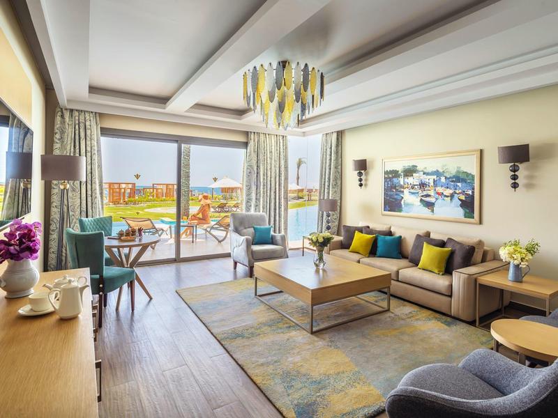 Bright hotel room with sofa, armchair, TV, and balcony with a view.