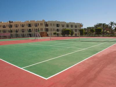 Leerer Tennisplatz mit grünem Spielfeld und rotem Untergrund vor hellen Gebäudezeilen unter blauem Himmel.