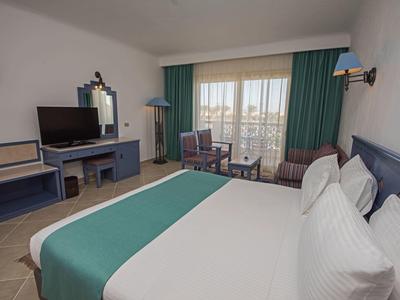 Helles Hotelzimmer mit großem Bett, Schreibtisch, Fernseher und Balkon mit Blick nach draußen.
