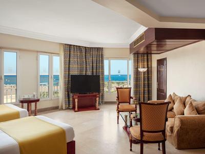 Prostorný hotelový pokoj se dvěma postelemi, pohovkou, židlemi a výhledem na moře skrz velká okna.