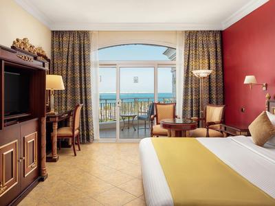 Hotelový pokoj s výhledem na moře, postel, skříň na TV, psací stůl a balkon s posezením.