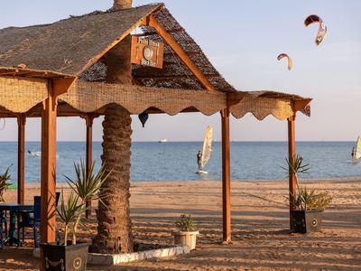 Playa con pabellón de madera, veleros y kitesurfistas en mar tranquilo al atardecer.