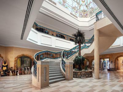 Prostorná hotelová hala s velkým schodištěm, palmou uprostřed a zdobeným stropem.