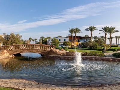 Malebná rezortní jezírko s malým mostem, fontánou, palmami a modrou oblohou.