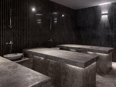 Moderní spa místnost s osvětlenými kamennými lavicemi a tmavými svislými dlaždicemi na stěnách.