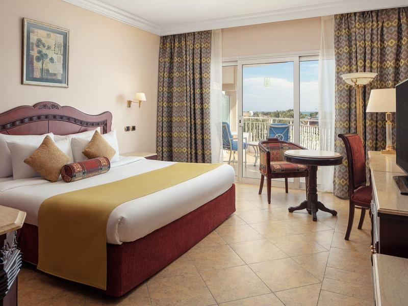 Hotelový pokoj s velkou postelí, dvěma židlemi, stolem a balkonem s výhledem na moře.
