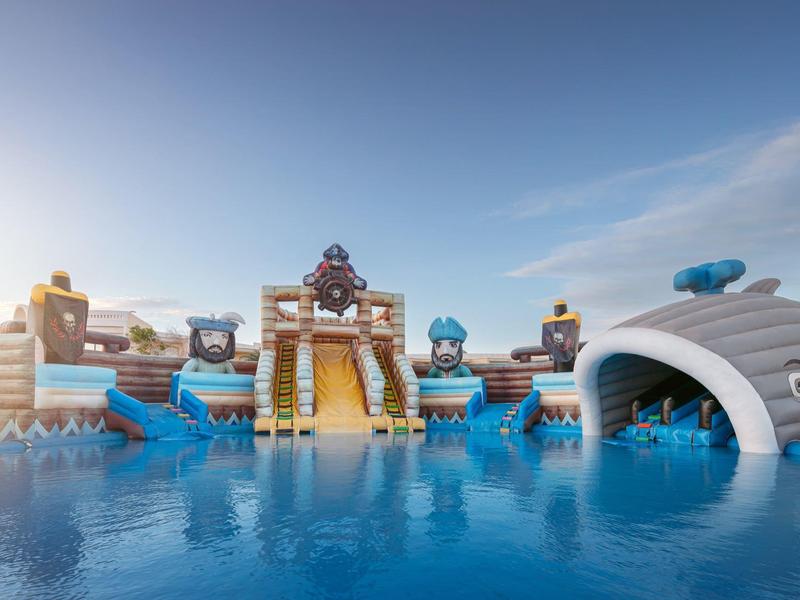 Barevné vodní hřiště s tobogány a konstrukcemi nad klidným bazénem pod jasnou oblohou.