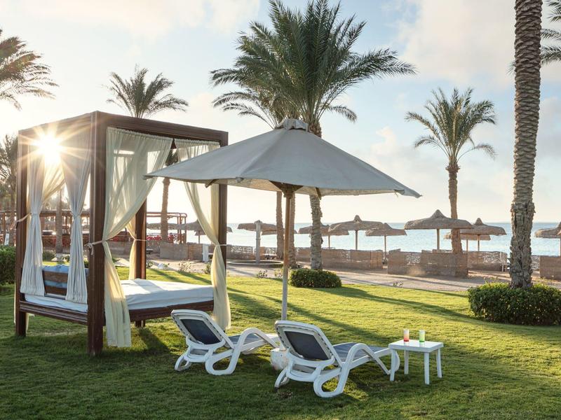 Slunečné plážové uspořádání s lehátky, baldachýnovou postelí a palmami na zelené trávě.