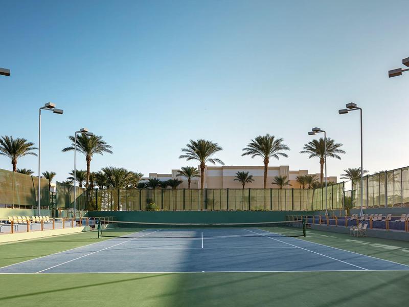 Slunečný venkovní tenisový kurt obklopený palmami a sedadly pod jasnou modrou oblohou.