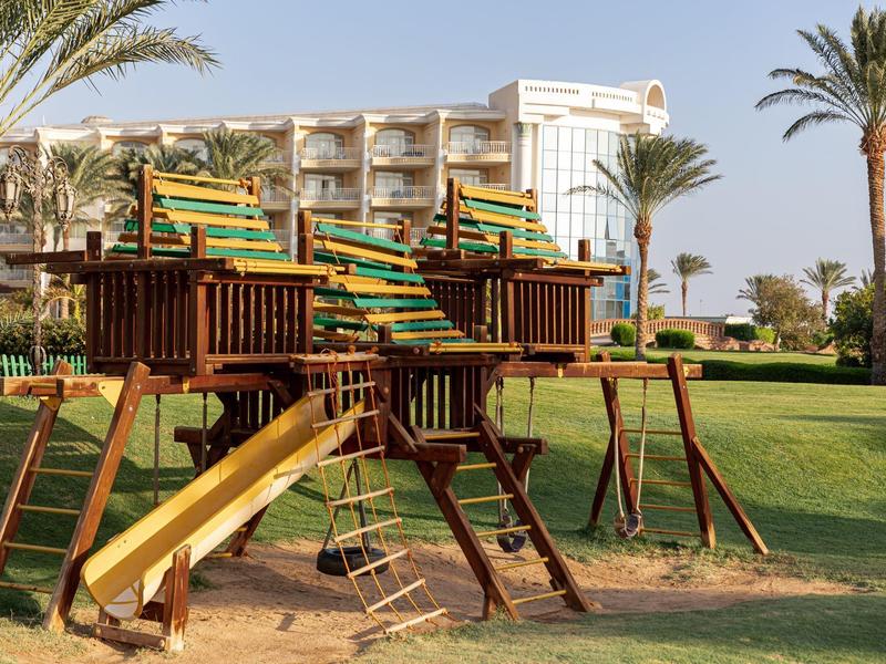 Dřevěné dětské hřiště s klouzačkami a houpačkami před hotelem s palmami.