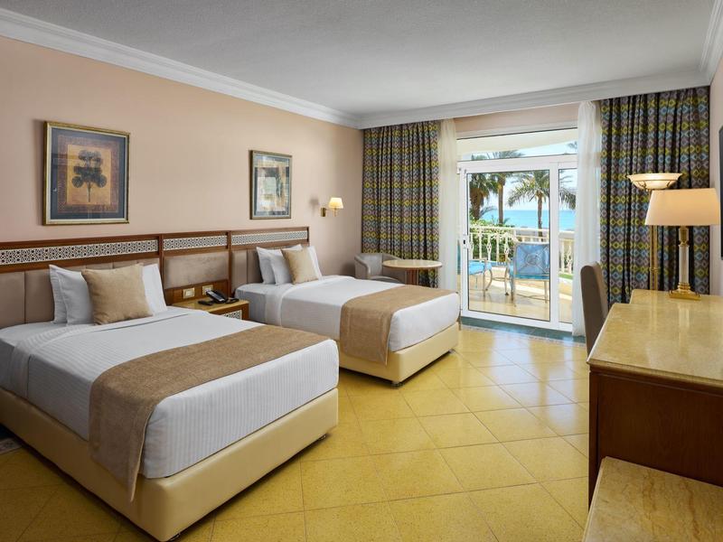 Hotelový pokoj se dvěma postelemi, dřevěným nábytkem a balkonem s výhledem na moře.