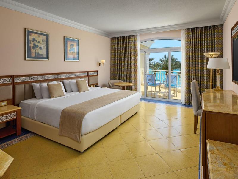 Hotelový pokoj s velkou postelí, balkonem s výhledem na palmy a moře, teplé osvětlení.