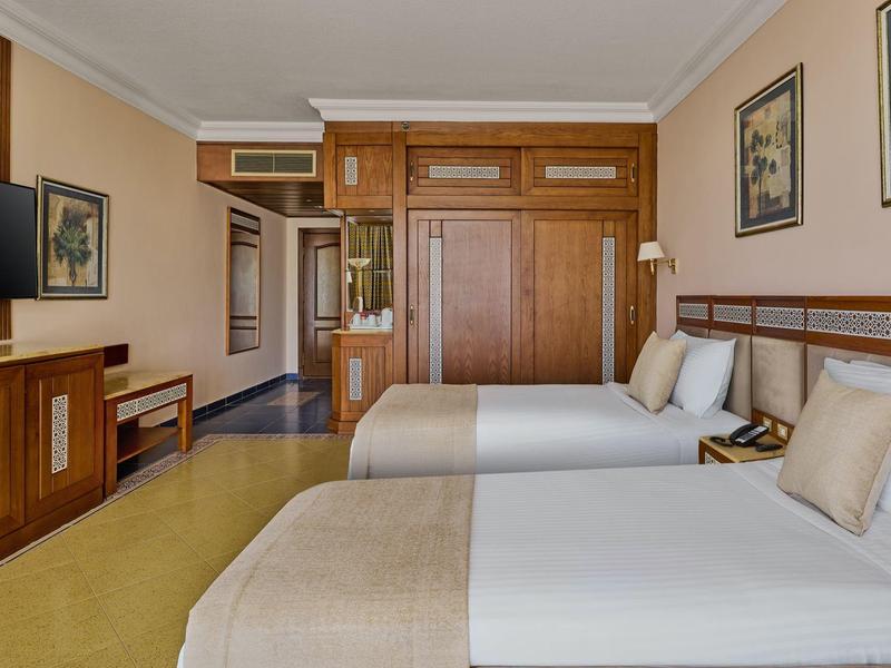 Hotelový pokoj se dvěma samostatnými postelemi, béžovým dekorem, televizí na stěně a dřevěným nábytkem.