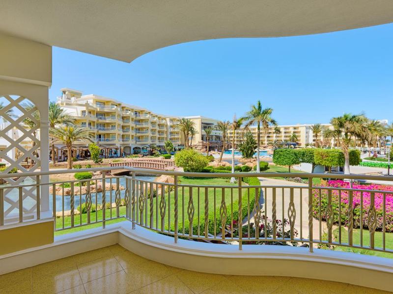 Výhled z balkonu na bazén, zahrady a budovy resortu pod jasnou oblohou.