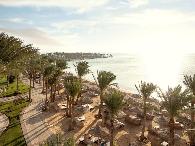 Slunečné plážové letovisko s palmami, lehátky a klidným mořem pod jasnou oblohou.