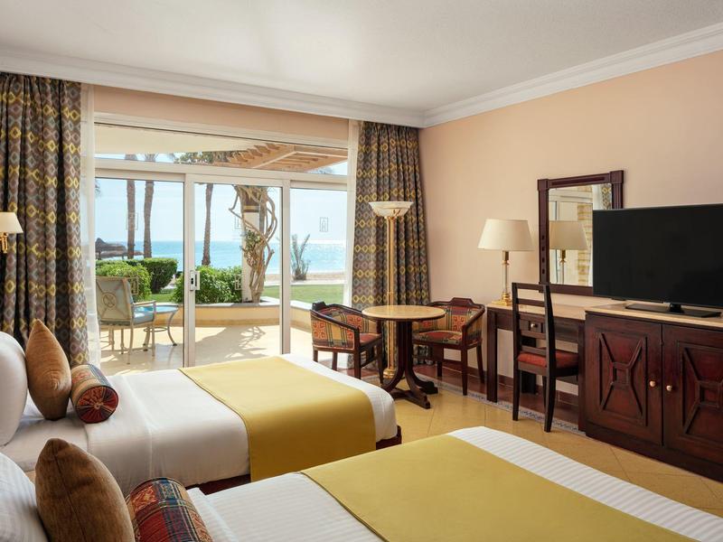 Hotelový pokoj se dvěma postelemi, žlutými přehozy, psacím stolem, TV a balkonem s výhledem na bazén a moře.
