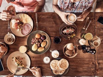 Mehrere Personen essen gemeinsam verschiedene Speisen auf einem hölzernen Tisch, darunter Brot, Fleisch und Getränke.
