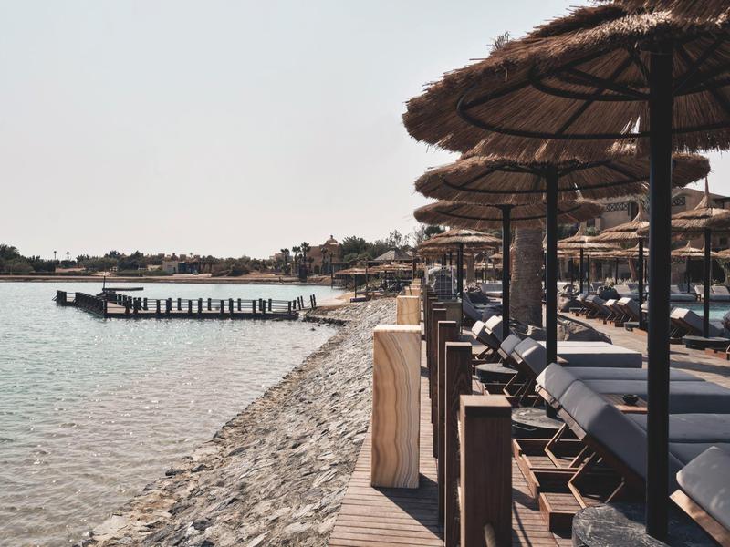 Trockene Landschaft mit Palmen, Sand und Felsen vor einer hellen Gebäudezeile unter klarem Himmel.