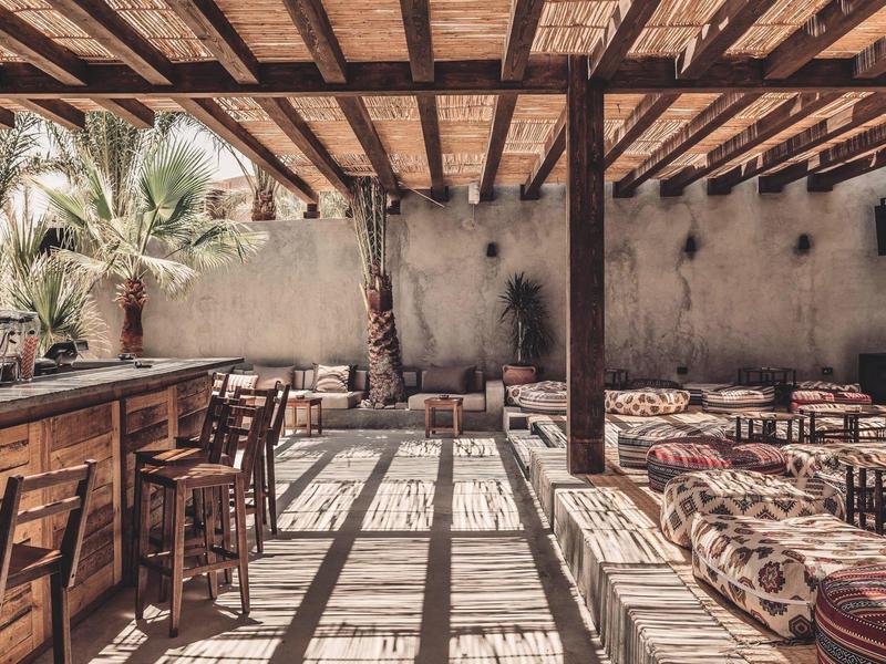Offener Raum mit Holzmöbeln, Sitzkissen, einer Bar und Palmen unter einer Holzdachkonstruktion.