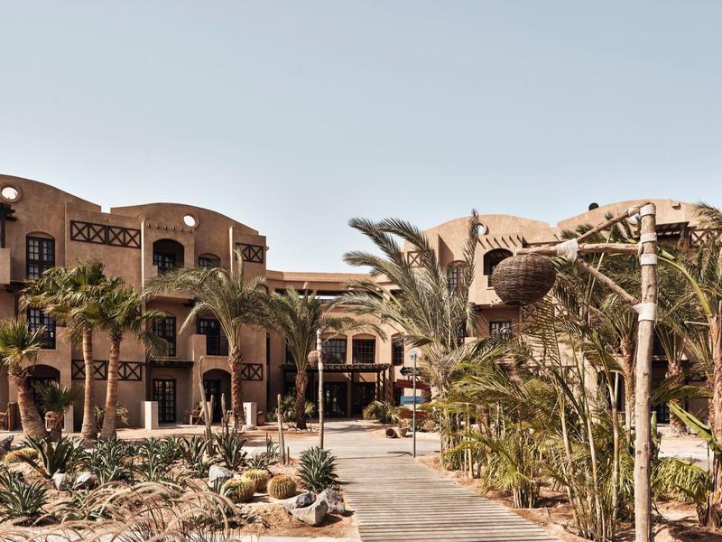 Große Anlage mit sandfarbenen Gebäuden, Palmen, Steinen und blauem Himmel im Hintergrund.