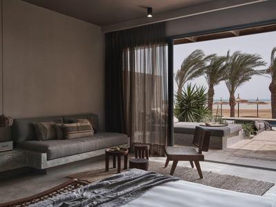 Modernes Schlafzimmer mit grauem Sofa, Holzmöbeln und Blick auf Terrasse mit Palmen und Meer.
