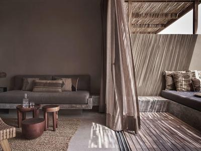 Moderner Sitzbereich mit grauem Sofa, Holztischen und offener Terrasse mit Liege und Vorhängen.