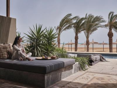Frau sitzt auf Terrasse mit Kissen, Palmen im Wind und Blick aufs Meer bei klarem Himmel.