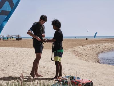 Zwei Männer stehen am Strand, einer mit Neoprenanzug, beide vor blauem Segel und Sand.