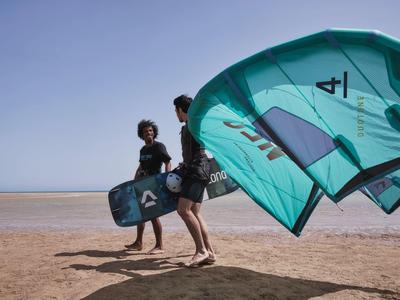Zwei Personen tragen Kitesurf-Ausrüstung am Sandstrand bei klarem Himmel.