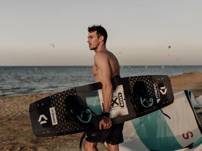 Ein Mann trägt an einem Strand ein schwarzes Kiteboard, im Hintergrund Meer und Himmel bei Sonnenuntergang.