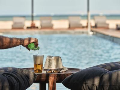 Person gießt Limette über zwei Biergläser neben Pool mit Liegen und Meer im Hintergrund.