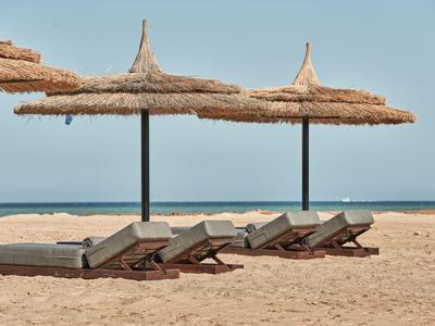 Sandstrand mit vier Liegen und zwei großen Strohmusterschirmen vor blauem Himmel und Meer.