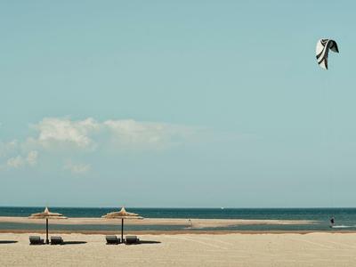 Leerer Sandstrand mit drei Sonnenschirmen, blauem Himmel und Drachen im Wind über dem Meer.