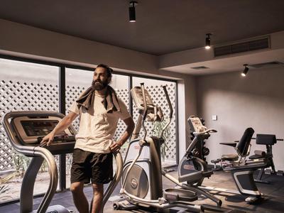 Mann mit Handtuch steht in modernem Fitnessraum mit Laufband und Ergometer, große Fenster im Hintergrund.