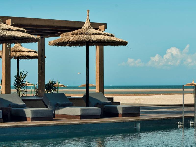 Pool mit Sonnenliegen und Strohdach am Strand bei klarem Himmel.