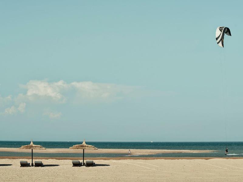 Weitläufiger Sandstrand mit Liegen, Sonnenschirmen und einem Drachen am Himmel.