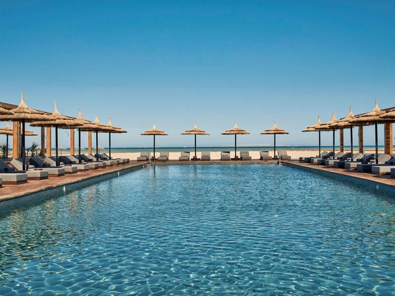 Großer Pool mit Sonnenliegen und Sonnenschirmen unter klarem blauem Himmel in einem Hotel.