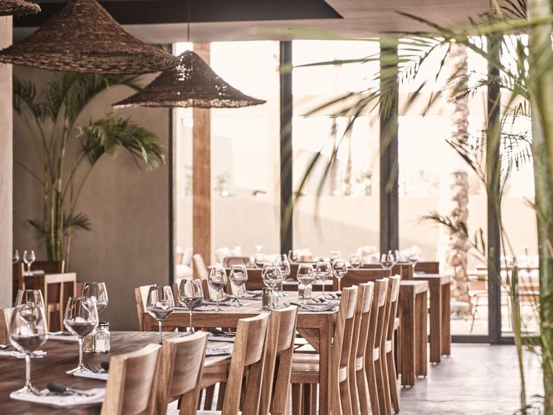 Helles, modernes Restaurant mit Holztischen und großen Fenstern, umgeben von Pflanzen.