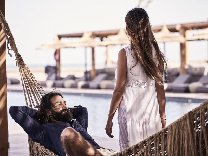 Entspannter Mann liegt in einer Hängematte, während eine Frau im weißen Kleid daneben steht.